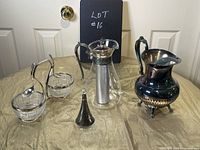 Photo shows assortment of silver plate and glass items on table including jug, condiment server, carafe, and small silver plate item