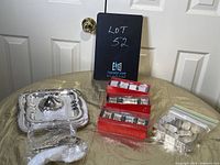 Disassembled tiered cake plate parts (three silver trays and central knob) displayed on the left with two red boxes containing silver napkin rings and extra napkin rings in plastic bag on the right.