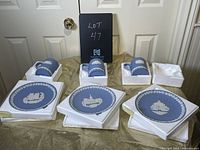 Three blue Wedgwood Christmas plates and three matching mugs in original white foam packaging, arranged on gold fabric.