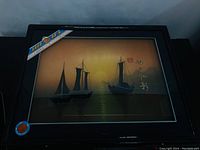 Full view of the framed wall art depicting sailing ships at sunset with Asian script, in a black wooden frame with glass covering.