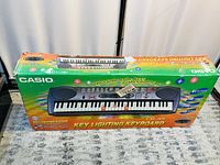 Photo of Casio LH-35 Key Lighting Keyboard boxed, showing packaging highlighting features like 61 keys, 100 tones, 100 rhythms, key lighting system, and included cable.