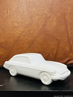 Side view of white ceramic car sculpture showing smooth matte finish and detailed molded lines on an orange-brown textured background.