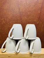 Five white ceramic cups arranged upside down on a wooden surface against a brown textured background. Three cups are on the bottom row and two on top.