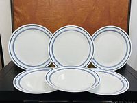 Four white ceramic dinner plates with blue double line around rim sitting against a brown background.