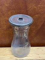 Front view of clear ribbed glass vase with lid on, showing hourglass shape and textured pattern.