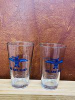 Pair of clear glass tumblers standing side by side on a wooden surface, both featuring a blue eagle graphic and the text 'WE ARE SHEP'.