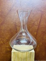 Photo showing the clear glass jar with a wide base and narrow neck on a light wood block against a brown background.