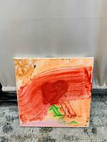 Front view of kid's canvas painting with predominantly red and orange colors, a large red heart in the center, and green and yellow accents, placed against a white curtain.