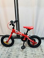 Red kids bike with black tires, red seat, black pedals, and yellow spoke reflectors shown from side angle on beige carpet with white curtain background.