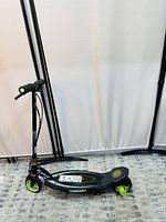 Full side view of the black and green Razor Power Core E90 scooter standing upright on a carpeted floor with a white backdrop, showing deck, wheels, handlebars, and kickstand.