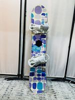 Front view of the snowboard showing colorful geometric pattern and attached white bindings.