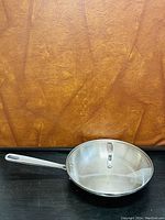 Same stainless steel frying sauté pan with tempered glass lid placed on top, shown against a brown textured background and black surface.