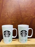 Two white ceramic Starbucks mugs with black siren logo and light green abstract circle and floral pattern, shown side by side on wooden platform with brown textured background.
