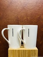 Two white tall Starbucks coffee mugs side by side from the front showing logo and handle.
