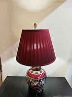 Front view of the vintage ginger jar table lamp showing maroon pleated shade and floral ceramic base with golden accents on dark background.