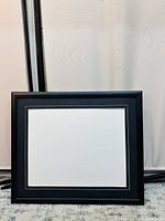 Black wooden picture frame with white mat placed on a carpet against a plain backdrop. Shows front view of the frame clearly.