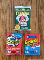 Front view of three sealed baseball card packs on wooden surface: one 1991 Score Series 1 and two Donruss Baseball Puzzle and Cards showing logos and graphics.