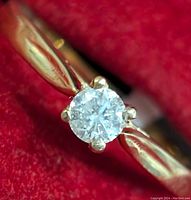 Close-up front view of a yellow gold ring with a round natural diamond in four-prong setting against a red background.