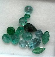 Top view photo of 14 loose genuine emerald gemstones in various shapes and sizes resting on white fabric background, showing their green hues and natural inclusions.