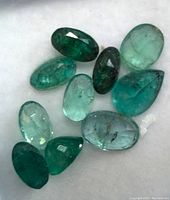 Top view of eleven assorted emerald gemstones showing different cuts and shades of green on white background.