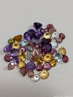 Collection of loose multi-colored faceted gemstones displayed on a white background, showing colors such as purple, yellow, pink, orange, green, and clear in various shapes and cuts.