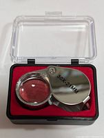 Close-up of metal jeweler's loupe showing folding design, magnification (30X), lens size (21mm), and protective plastic case with red interior.
