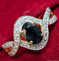 Close-up photo of a sterling silver ring with an oval deep blue sapphire center stone set in yellow gold-tone prongs, featuring a twisting band design encrusted with small round clear cubic zirconia stones.