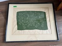 Full view of framed print labeled with green tape '644', showing white line drawings on green background depicting Indigenous figures and animals.