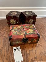 Three Asian-themed decorative boxes on wooden floor; one large red lacquered jewelry box with artwork and two smaller dark wood square boxes with intricate circular designs