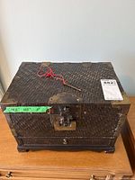 Front view of Asian styled wicker chest with visible metal locks, corner brackets, key tied with red string, and drawer at bottom.