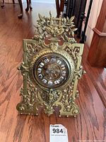 Full front view of ornate brass clock with floral motifs and urn decoration on top, Roman numeral dial visible.