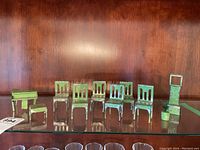 Front view of seven small tin chairs and one shorter piece with a mini potted plant, all with green paint worn with age on a glass shelf.