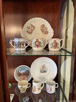 Display of 6 mugs and 3 plates with various English Coronation images on a glass shelf inside a cabinet. Plates include shell-shaped and round with royal portraits. Mugs include different styles with detailed royal crests and monarch images.