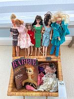 Six dolls displayed upright in a basket with a Barbie 35th Anniversary booklet and various doll clothes at the bottom of the basket.