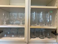 Cabinet shelves with 5 snifters on top left, 8 water glasses (6 smaller and 2 taller) on top right, 7 highball glasses on lower left, 4 juice glasses and 1 shot glass on lower right.