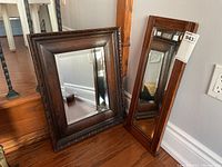 Two wood framed mirrors positioned on floor by stairs, one larger with decorative carved edge and one smaller with plain wood frame