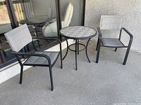 Photo showing two black metal frame patio chairs with gray fabric seats and backs alongside a round table with ceramic top on a patio floor.