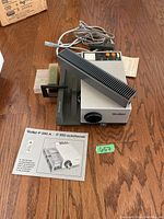 Rollei P350 A projector with power cords, slide tray, vent cover, and instruction manual on wooden floor.