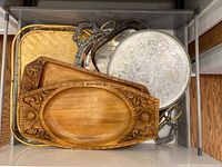 Photo showing a top-down view of assorted serving trays including wooden trays with carved detailing, metal trays with handles, and a bamboo tray.