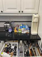 Wide view of countertop showing SodaStream unit, toaster, clear travel cups, and boxes of food storage products. Two drawers below counter with kitchen utensils visible.