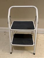 Front view of a white 2-step metal stool with black non-slip step surfaces and a top handle for support.
