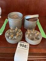 Set of 10 clear Swarovski crystal name holders with gold-tone loops placed in two grey metal storage boxes with foam inserts, displayed on wooden surface.