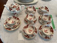 Teapot, 7 teacups with saucers, and 4 small plates arranged on a tablecloth showing floral and ornate maroon pattern with gold accents.