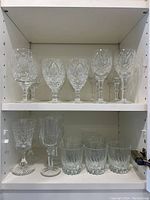 Full shelf view of assorted stemmed wine glasses and tumblers, showing 14 glasses (top and bottom shelves) with cut glass and plain clear styles.