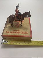 Front-right angle of metal figure seated on horse with measuring tape showing approximately 4 inch length, figure placed on original box with water damage.