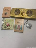 Photo of three tiny books and a Royal souvenir tin box with portraits, plus a tin medal with a safety pin.