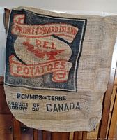 Photo showing large burlap potato sack with printed 'Prince Edward Island PEI Potatoes' text, a red map of PEI, and 'Product of Canada' text.