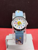 Front facing image of girls quartz watch with blue silicone band featuring My Little Pony pony and star designs and white dial with multicolored numbers and yellow horseshoe motif.
