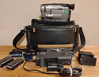 Complete Sony Handycam video camera package showing camera on top of soft carrying case and accessories including batteries, charger, cables, battery adapter, and shoulder strap arranged in front