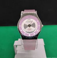Front view of women's translucent pink band quartz watch on stand against green and black background.